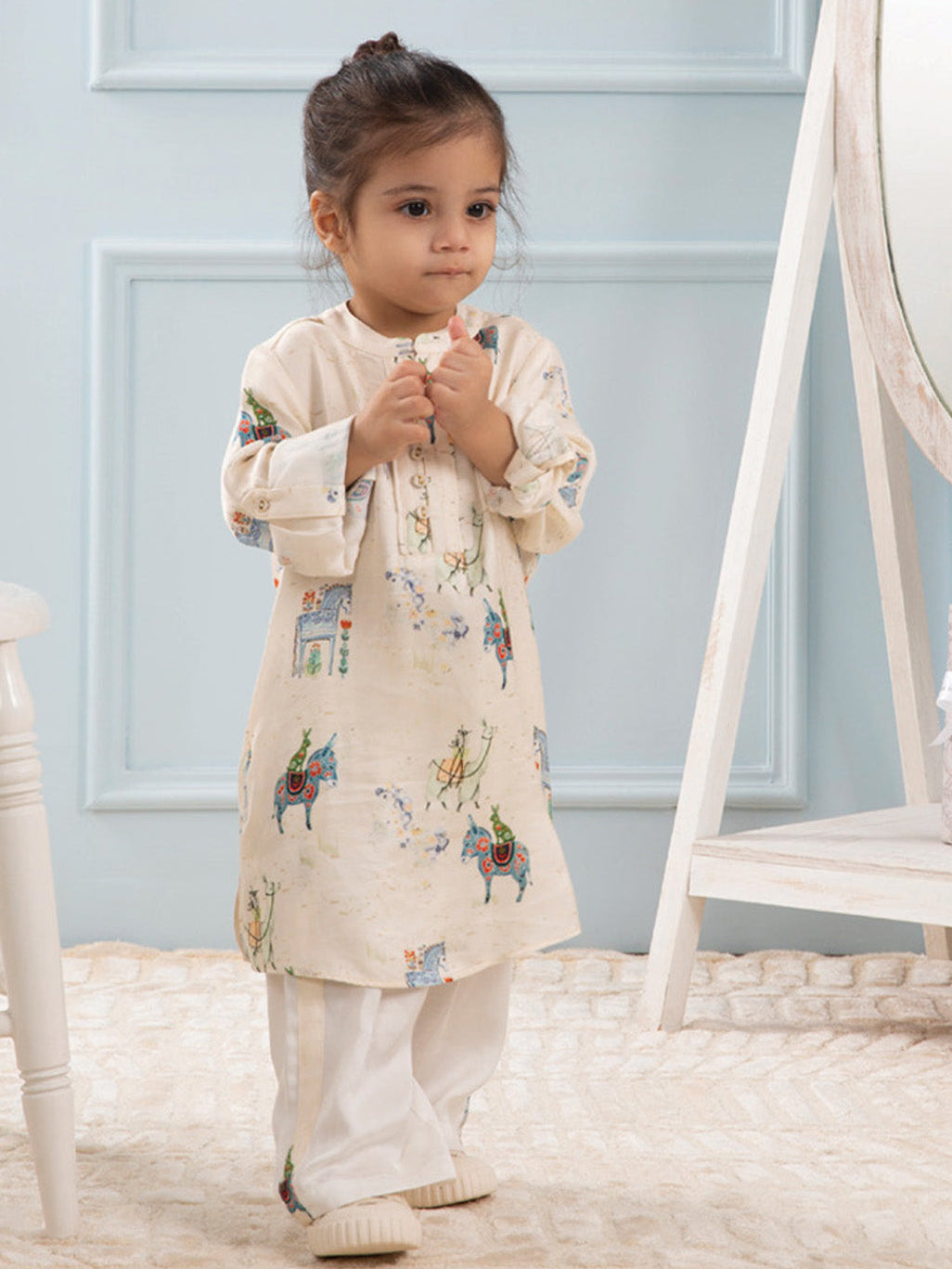A young boy wearing a beige and white printed co-ord set with hand-painted designs, standing in a room with light blue walls.
