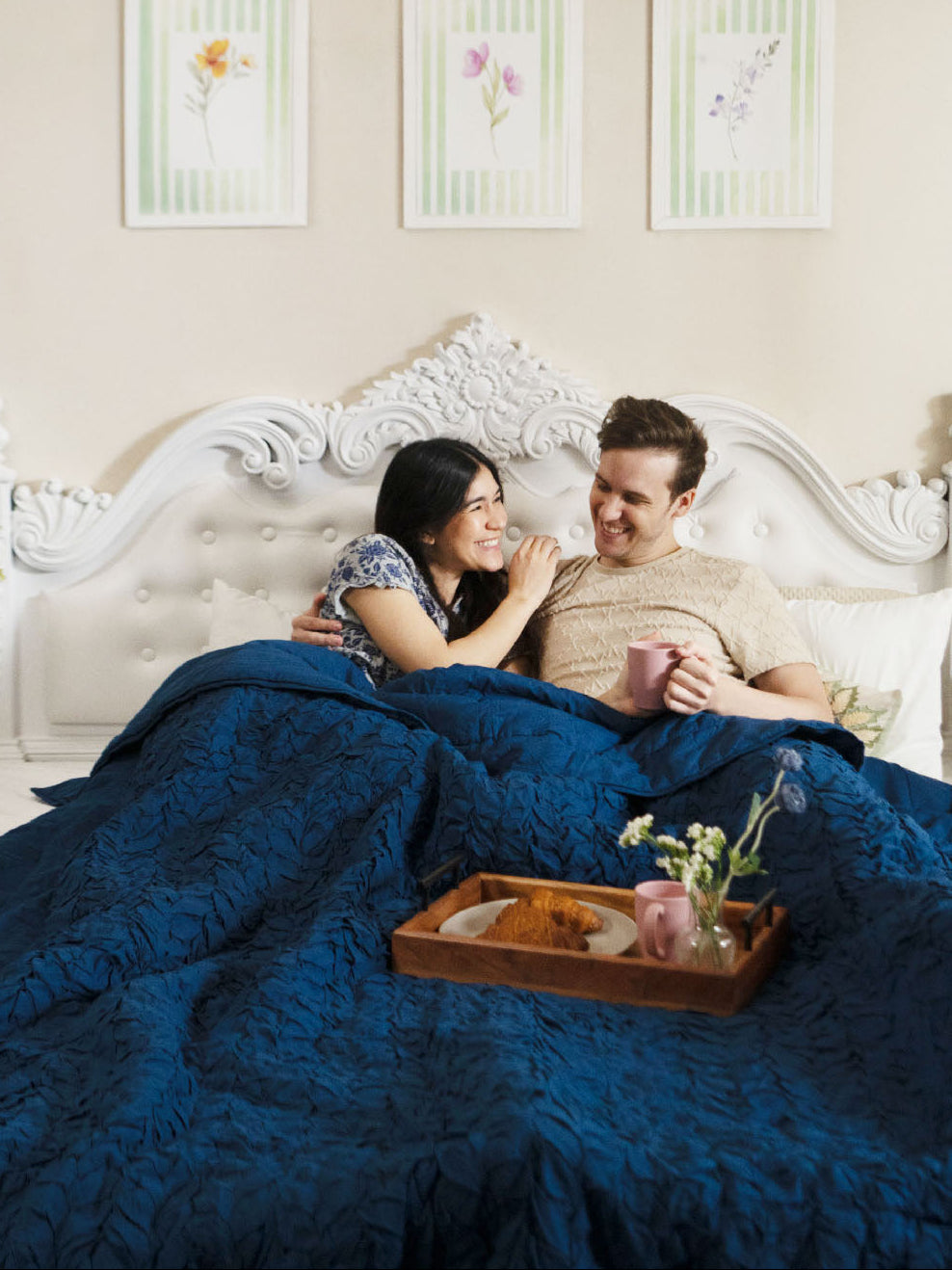 Couple lying in bed under a blue comforter with a tray of flowers and candles.
